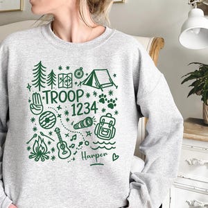May include: A light gray sweatshirt with a green camping-themed graphic. The design includes trees, a tent, a map, a compass, a guitar, a campfire, and the text "TROOP 1234". The name "Harper" is also included.