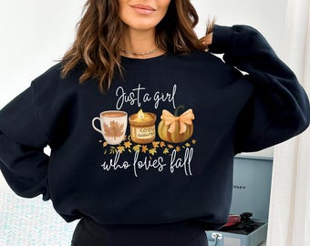 May include: Black sweatshirt with a fall-themed graphic. The design includes a coffee mug with a maple leaf, a candle, and a pumpkin with a bow. The text reads "Just a girl who loves fall."