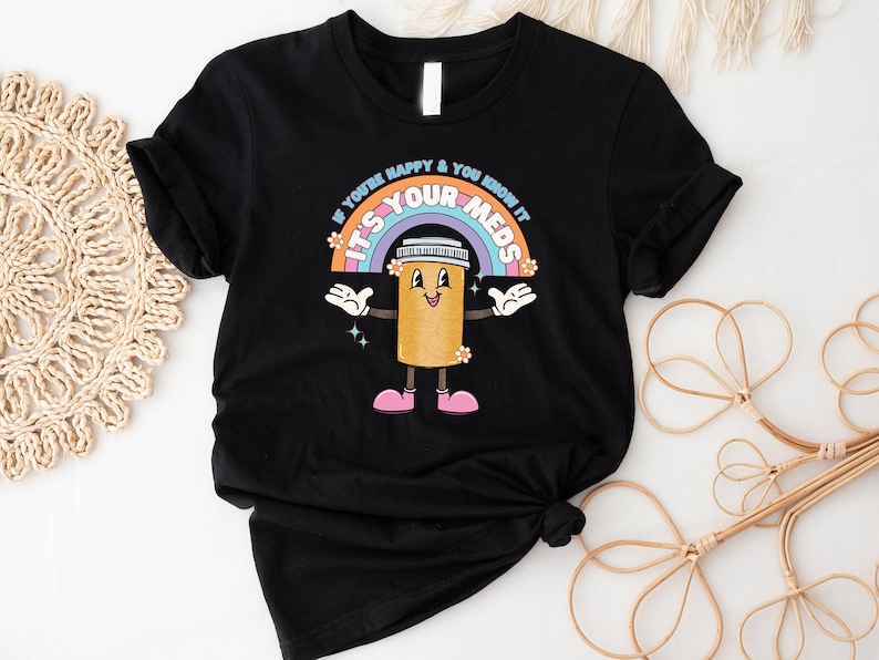 Peut inclure: T-shirt noir avec un arc-en-ciel et une pilule de dessin anim&eacute; avec le texte "Si vous &ecirc;tes heureux et que vous le savez, c'est vos m&eacute;dicaments".
