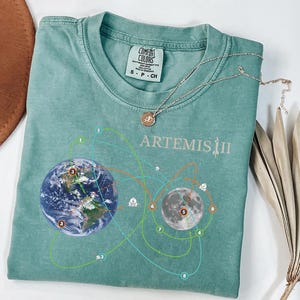 May include: A teal Comfort Colors t-shirt featuring a graphic of Earth and the Moon with connecting lines and the text "ARTEMIS II". A silver necklace with a circular pendant rests on the shirt. A dried plant is next to the shirt.