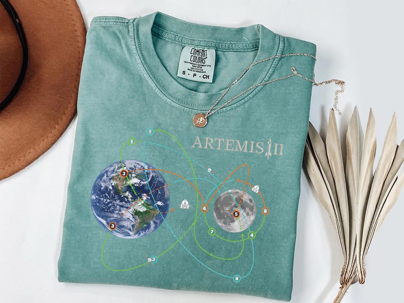 May include: A teal Comfort Colors t-shirt featuring a graphic of Earth and the Moon with connecting lines and the text "ARTEMIS II". A silver necklace with a circular pendant rests on the shirt. A dried plant is next to the shirt.