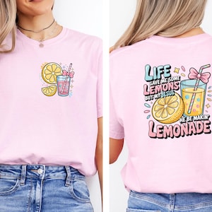 May include: Light pink t-shirt with a colorful lemonade graphic on the front and back. The front features a lemon slice, a drink, and a bow. The back says "Life gave me some lemons but my Jesus he be makin' lemonade."