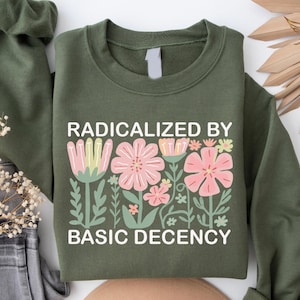May include: Olive green sweatshirt with a floral design and the text "RADICALIZED BY BASIC DECENCY" in white. The floral design features pink and yellow flowers with green leaves.