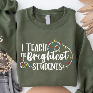 May include: Olive green sweatshirt with the text "I Teach the Brightest STUDENTS" in white, encircled by a string of colorful Christmas lights. The sweatshirt is folded, with a portion of the sleeve visible.