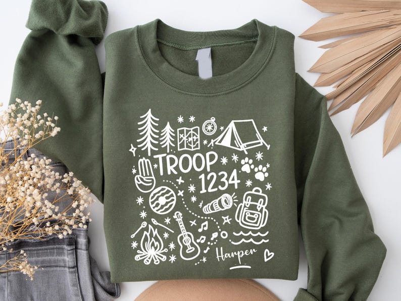 May include: Olive green sweatshirt with white graphic of camping elements, including trees, tent, map, and guitar. Text reads "TROOP 1234" and "Harper". The sweatshirt is folded, with a portion of the sleeve visible.