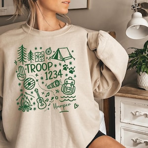 May include: Beige sweatshirt with a green graphic design featuring camping-themed illustrations. The design includes trees, a tent, a compass, and the text "TROOP 1234". The name "Harper" is also included.