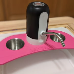 May include: A pink water bottle dispenser with a black and white pump and two stainless steel cup holders. The dispenser is on a white surface with the word "JAYDEN" printed on the front.