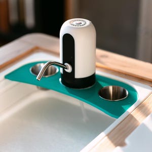 May include: A white and black electric water dispenser with a silver spout, sitting on a teal blue tray with two stainless steel cups. The dispenser is on a wooden surface.