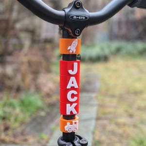May include: A red, white, and orange personalized handlebar name tag for a child's bike. The name "JACK" is spelled out in white letters on the red portion of the tag. The tag also features a rocket ship and a train design.