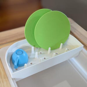May include: A white plastic dish drying rack with a blue cup and two green plates. The rack has a curved shape and multiple holes for drainage.