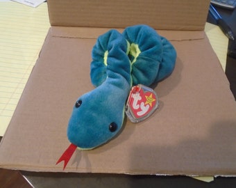Pode incluir: Um brinquedo de pelúcia de cobra turquesa com olhos de botão pretos e uma língua vermelha. A cobra tem um detalhe amarelo e uma etiqueta Ty anexada. O brinquedo está em uma superfície de papelão marrom.