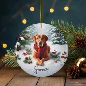 May include: A round ceramic Christmas ornament featuring a brown dog wearing a red plaid scarf, lying in the snow. The ornament has a winter scene with pine trees and berries. The name "Gunner" is written on the ornament.