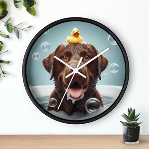 May include: A brown Labrador Retriever dog with a yellow rubber duck on its head is in a bathtub full of bubbles. The dog is smiling and has its tongue sticking out. The image is on a round clock with black hands and a black frame.