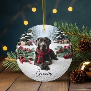 May include: Round ceramic Christmas ornament featuring a black Labrador Retriever wearing a red and black plaid scarf, lying in a snowy winter scene. The dog's name, "Gunner", is written in cursive. The ornament has a gold string for hanging.