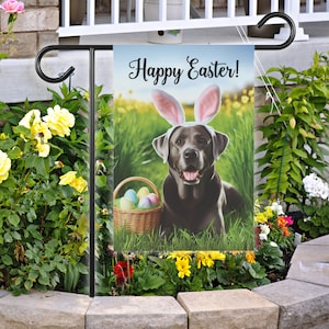 May include: A black Labrador Retriever dog wearing bunny ears and a basket of Easter eggs. The dog is sitting in a grassy field with yellow flowers. The text "Happy Easter!" is printed on the flag.