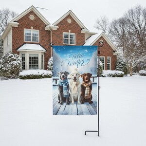 May include: A winter-themed garden flag featuring three Labrador Retrievers wearing scarves and hats, with the words "Hello Winter" in script. The dogs are sitting on a snowy wooden deck, with a winter landscape background.