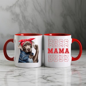 Puede incluir: Dos tazas de cer&aacute;mica blanca con bordes rojos. Una taza presenta un perro marr&oacute;n y gris con un pa&ntilde;uelo rojo y una chaqueta vaquera con el pu&ntilde;o en alto. La otra taza tiene el texto "BOSS MAMA BOSS" repetido en rojo.
