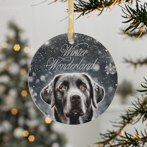 May include: Round Christmas ornament featuring a black Labrador Retriever with brown eyes, set against a snowy background. The text "Winter Wonderland" is written in elegant script. The ornament has a gold ribbon for hanging.