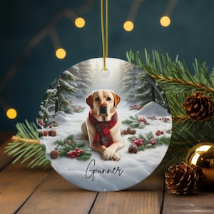 May include: Round ceramic ornament featuring a yellow Labrador Retriever wearing a red plaid scarf, lying in a snowy forest scene. The dog's name, "Gunner," is written in script. The ornament hangs from a gold string.