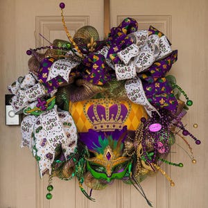 May include: A festive Mardi Gras wreath with a crown, mask, and ribbons. The wreath features purple, green, gold, and white accents, including beads and the text "Mardi Gras" on the ribbons. The wreath is designed to hang on a door.