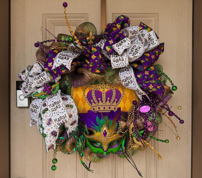 May include: A festive Mardi Gras wreath with a crown, mask, and ribbons. The wreath features purple, green, gold, and white accents, including beads and the text "Mardi Gras" on the ribbons. The wreath is designed to hang on a door.