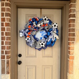 May include: A decorative wreath with a soccer theme, featuring soccer balls, musical notes, and blue and orange ribbons. The wreath is hanging on a beige door, with a brick and stone exterior.