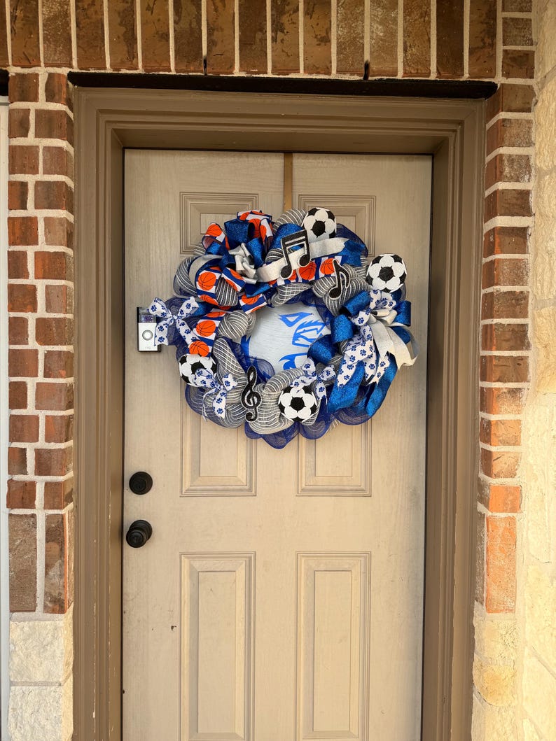 May include: A decorative wreath with a soccer theme, featuring soccer balls, musical notes, and blue and orange ribbons. The wreath is hanging on a beige door, with a brick and stone exterior.