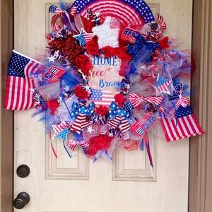 May include: A patriotic wreath with red, white, and blue ribbons, stars, and flowers. The wreath includes small American flags and a sign that reads "Home of the Free Because of the Brave."