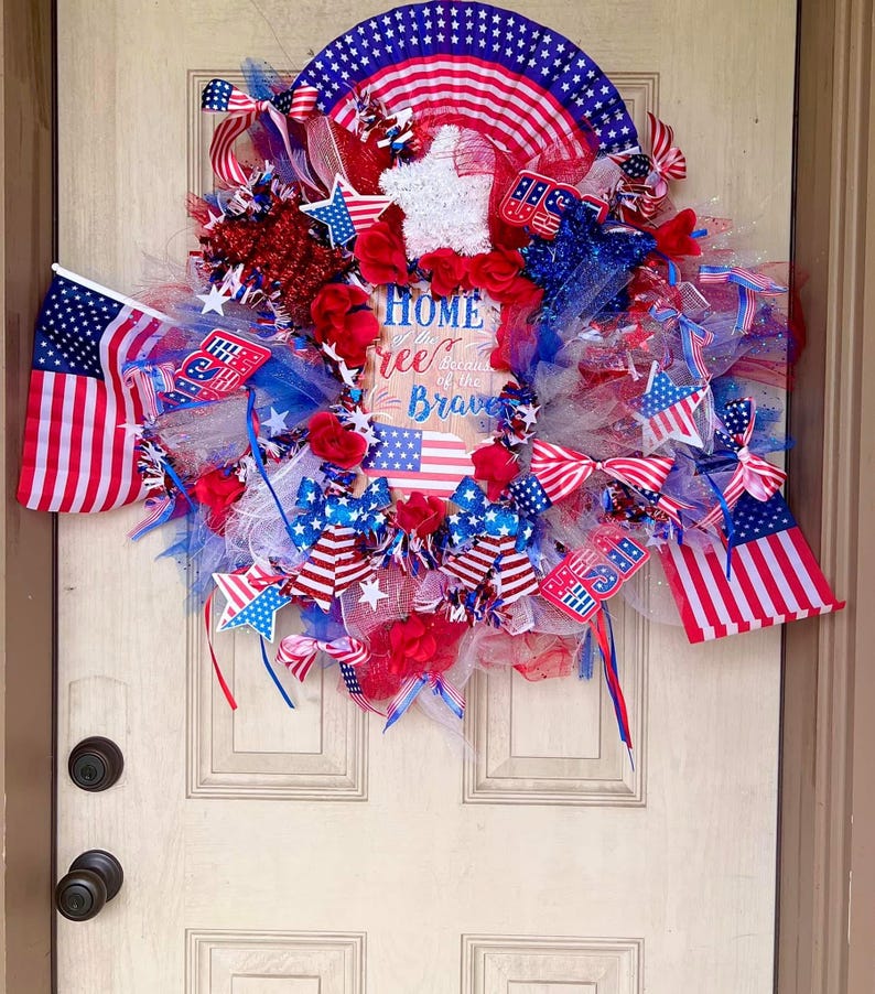 May include: A patriotic wreath with red, white, and blue ribbons, stars, and flowers. The wreath includes small American flags and a sign that reads "Home of the Free Because of the Brave."