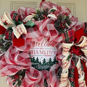 May include: A festive Christmas wreath with red and white mesh, plaid ribbons, and decorative accents. The wreath features a sign that reads "Welcome to the Hamlin's home" and "Merry Christmas". Includes pinecones and greenery.