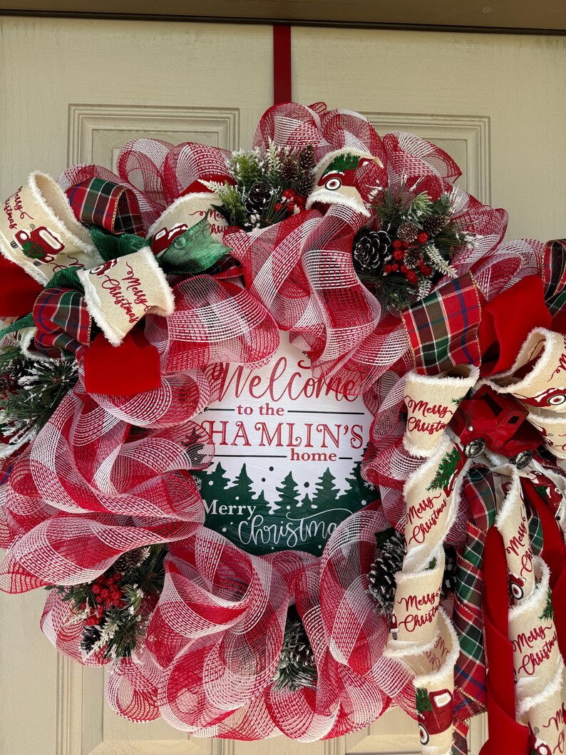 May include: A festive Christmas wreath with red and white mesh, plaid ribbons, and decorative accents. The wreath features a sign that reads "Welcome to the Hamlin's home" and "Merry Christmas". Includes pinecones and greenery.