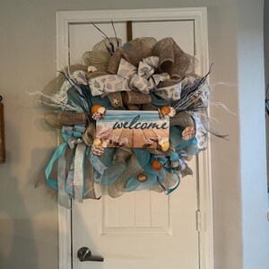May include: A decorative beach-themed wreath with a burlap base, adorned with seashells, blue ribbons, and a sign that says "welcome." The wreath is designed to hang on a door and has a coastal aesthetic.