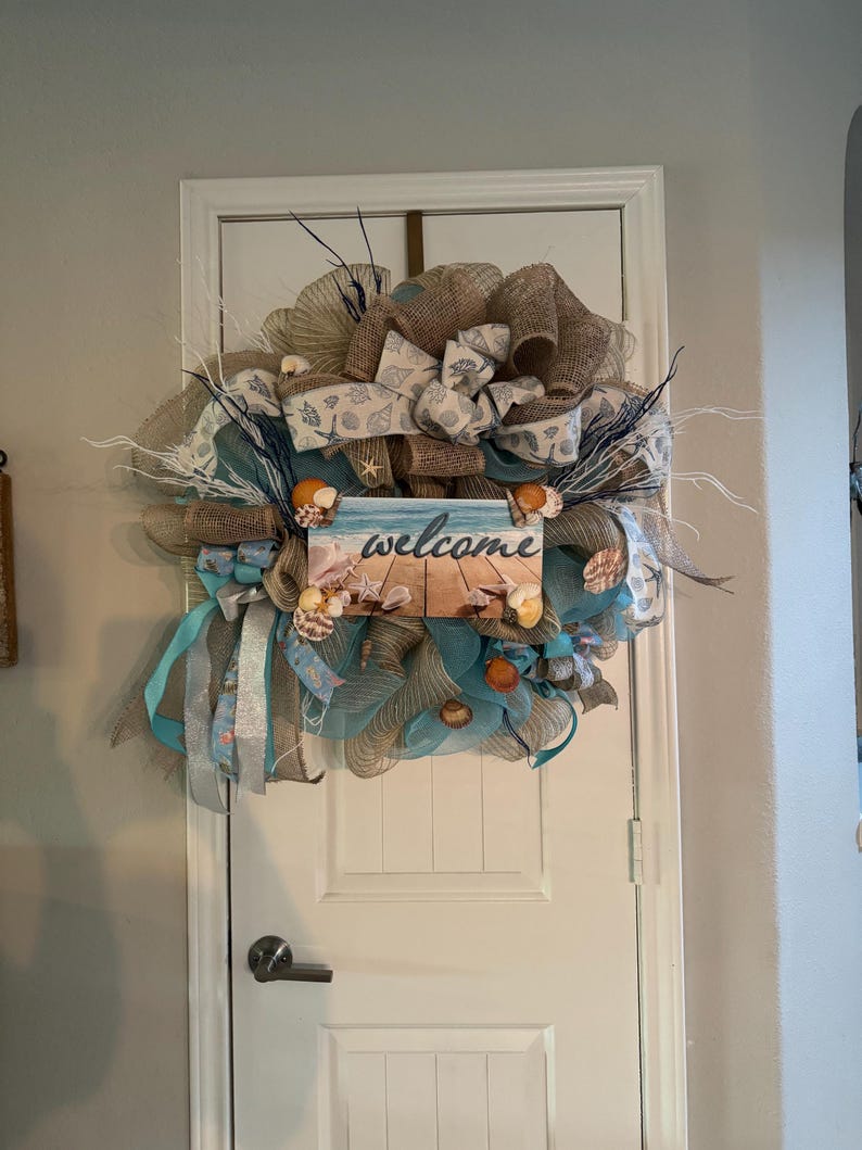 May include: A decorative beach-themed wreath with a burlap base, adorned with seashells, blue ribbons, and a sign that says "welcome." The wreath is designed to hang on a door and has a coastal aesthetic.