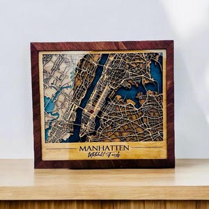 May include: A wooden map of Manhattan, New York City, with a dark wood frame. The map is laser cut and shows the streets and waterways of the city. The map is personalized with the text "Manhattan" and "Withall Family".