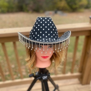 May include: A black cowboy hat with a rhinestone design and fringe. The hat is on a mannequin head.
