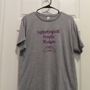 Könnte beinhalten: Ein graues T-Shirt mit dem lila Schriftzug "Pathological People Pleaser" und einem lila Herzsymbol aus Händen. Das T-Shirt hängt an einem schwarzen Kleiderbügel vor einer weißen Tür.