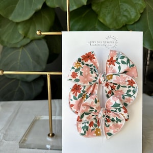 May include: Two floral print hair bows with pink, red, and green colors. The bows are displayed on a white card with the text "Happy Day Designs" and "Handmade with love".