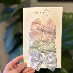 May include: A set of three hair bows in pink, green, and blue with floral patterns. The bows are displayed on a white card with the text "Happy Day Designs Kansas City, MO".