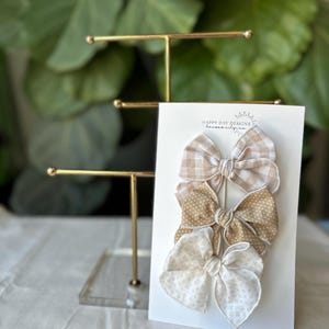May include: Three hair bows on a white card with the text "Happy Day Designs" in black. The bows are made of beige and white fabric with different patterns. The top bow is a beige and white gingham pattern, the middle bow is a beige polka dot pattern, and the bottom bow is a white floral pattern.