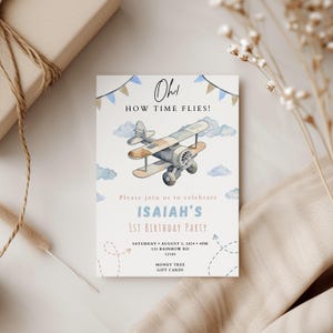 Puede incluir: Un avión azul y marrón con nubes y pancartas en el fondo. El texto de la invitación dice "Oh! How Time Flies!" y "Please join us to celebrate Isaiah's 1st Birthday Party". La fecha y hora de la fiesta se indican como "Saturday August 3, 2024 4PM" y la dirección es "123 Rainbow Rd 12345". La invitación también incluye la información "Money Tree Gift Cards".