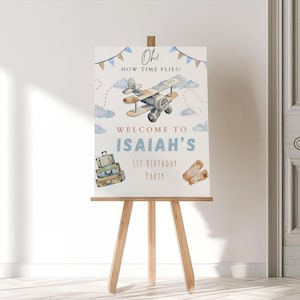 May include: A white welcome sign with blue and brown accents. The sign features a cartoon airplane, luggage, and the text "Oh! How time flies! Welcome to Isaiah's 1st Birthday Party."