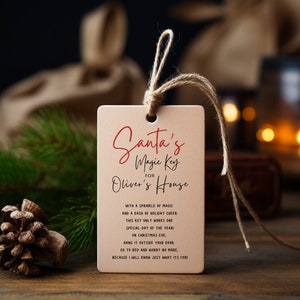 Op de afbeelding: Een bruin papieren label met een rood touwtje eraan. Het label heeft de tekst "Santa's Magic Key for Oliver's House" met een bericht over een speciale sleutel die alleen op kerstavond werkt.