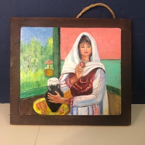 May include: Oil painting of a woman in traditional attire holding a dark vase and a red fruit. The painting is set in a framed wooden panel with a rope for hanging. The background features a window with a green landscape and a pink wall.