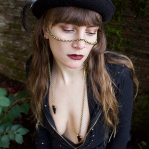 May include: A woman wearing a black hat with a feather, a black jacket, and a gold chain necklace with a black pendant. She has a nose ring and a lip ring.