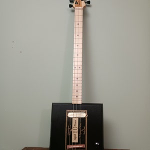 May include: A handmade black cigar box guitar with a wooden neck and headstock. The headstock has black tuning pegs and the brand name "Wave Part" is visible. The box features a label that reads "Aged 10 Years".