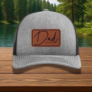 May include: Gray trucker hat with a black mesh back and a curved brim. The hat features a brown leather patch with the word "Dad" in a cursive font and the text "est. 2025".