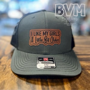May include: A dark gray and black trucker hat with a brown leather patch that reads "I LIKE MY GIRLS A LITTLE BIT OLDER". The hat is made by Richardson and has a 112 Trucker label.