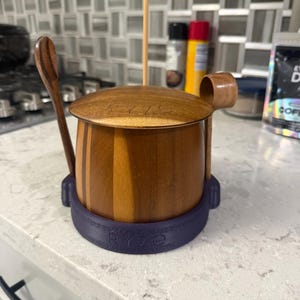 Può includere: Un vasetto di miele in legno con coperchio e un cucchiaino di legno. Il vaso è cilindrico con una base viola scuro con la scritta "RYZE". Un misurino di miele in legno è in posizione verticale. Il vaso è su un piano di lavoro bianco.
