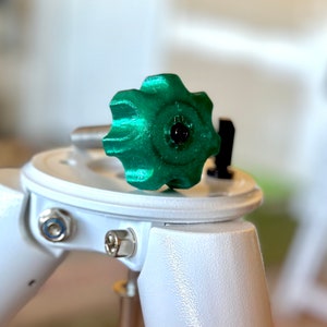 Może przedstawiać: Zielony plastikowy pokrętło w kształcie gwiazdy jest przymocowane do białego statywu. Pokrętło służy do regulacji wysokości statywu.