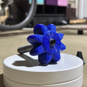 May include: A blue plastic gear with a threaded shaft, resting on a white circular base. The gear has a starburst design with eight points.